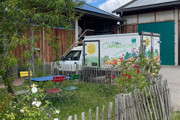 KI generiert: Ein Lieferwagen von "Biohof Appfelbacher" steht vor einem Gebäude; daneben ein bunter Sitzbereich im Freien.