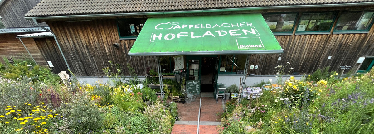 KI generiert: Ein Hofladen mit grünem Vordach, umgeben von blühenden Pflanzen. Text: "Apfelbacher Hofladen" und "Bioland".