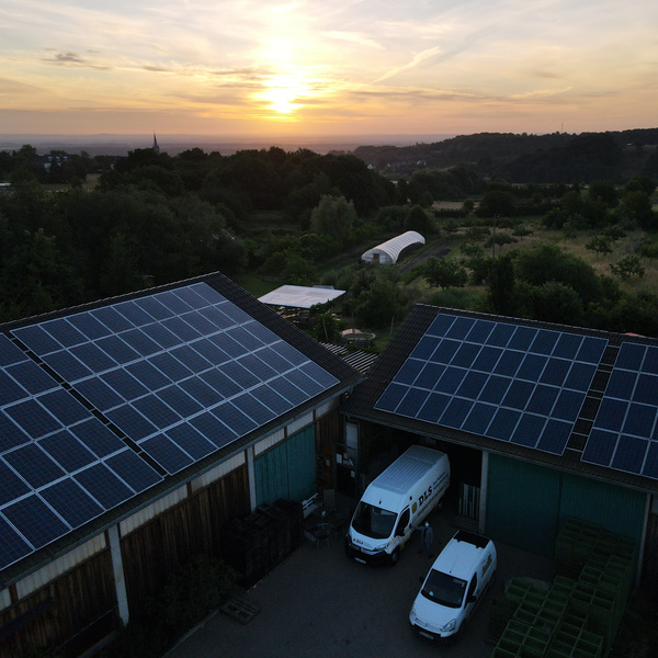 KI generiert: Gebäude mit Solarpanels auf den Dächern bei Sonnenuntergang, ländliche Umgebung mit Gewächshaus.