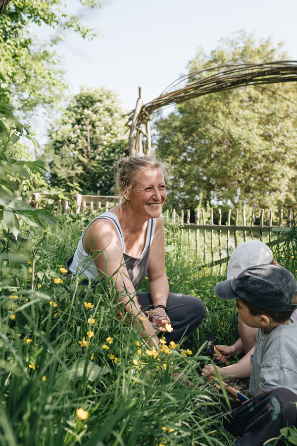 KI generiert: Eine Frau und zwei Kinder sitzen lächelnd im Gras, umgeben von gelben Blumen und einem Holzzaun.