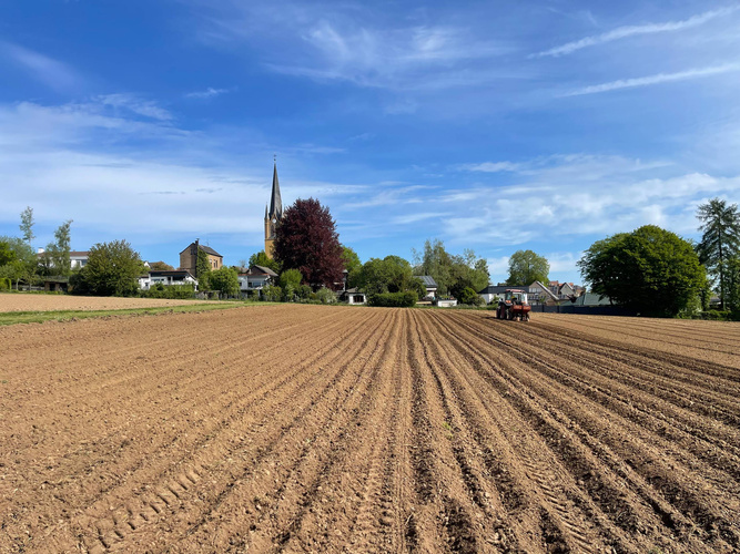 KI generiert: Ackerfeld mit Traktor, in der Ferne ein Kirchturm und einige Häuser, bewölkter blauer Himmel.