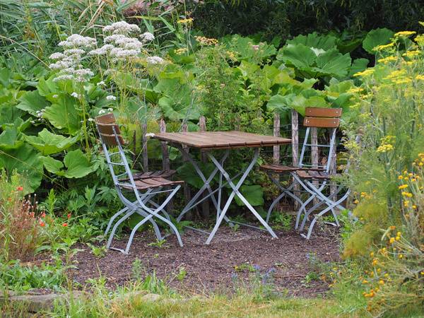 KI generiert: Ein Holztisch mit zwei Stühlen in einem üppigen Garten voller grüner Pflanzen und blühenden Blumen.