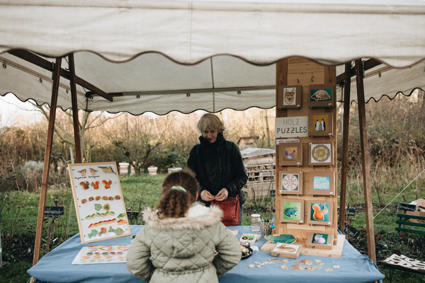 KI-generierte Beschreibung: Holzpuzzle-Stand mit Verkäuferin und Kundin. Text: "HOLZ PUZZLES".