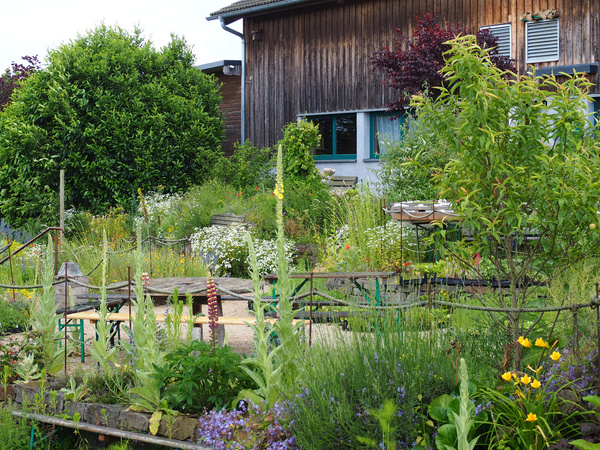 KI generiert: Ein üppiger, blühender Garten vor einem Holzhaus mit Bänken und Tischen.