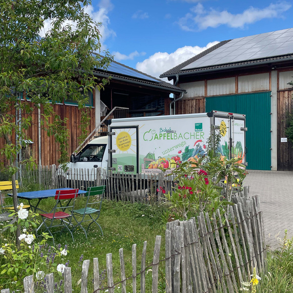 KI generiert: Ein Lieferwagen von "Biohof Apfelbacher" steht vor einem Hof, daneben bunte Gartentische und Bänke.