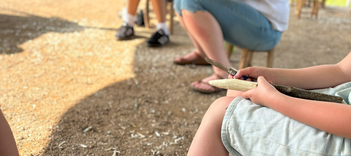 KI generiert: Ein Kind schnitzt draußen mit einem Messer an einem Stock. Mehrere Menschen sind im Hintergrund sichtbar.