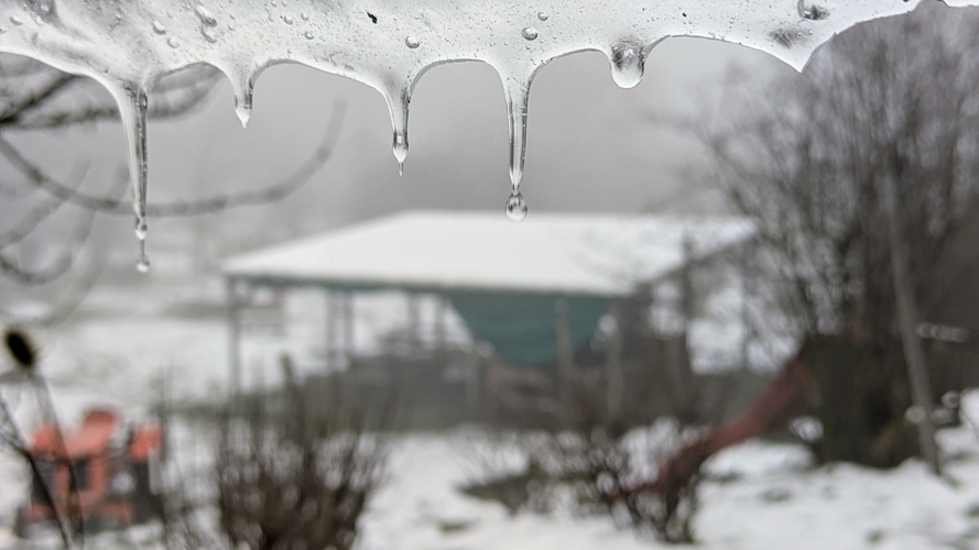KI generiert: Eiszapfen im Vordergrund, unscharfe verschneite Landschaft mit Gebäude im Hintergrund.